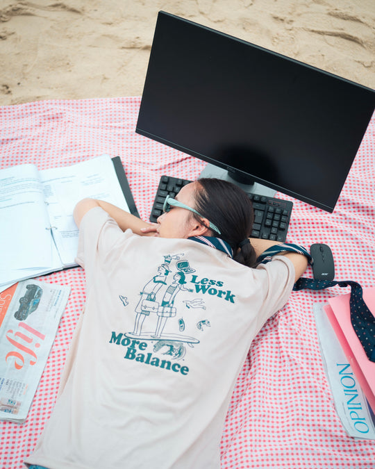 woman falling asleep by a computer on the beach wearing a graphic t shirt which says work life balance