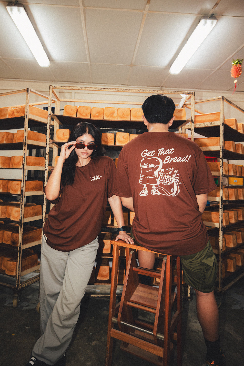 man's back and woman front facing a camera in brown graphic t shirt in a bakery